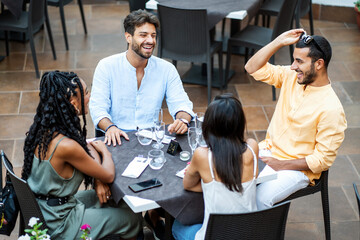 Diverse friends enjoying outdoor dining: - Multicultural group laughing at restaurant terrace, young adults socializing, summer lifestyle, inclusive friendship