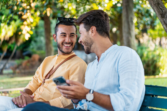Diverse friends sharing smartphone outdoors: Gay couple enjoying technology in a park - LGBTQ relationship, millennial men using mobile app together