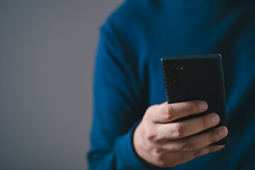 Person holding smartphone in hand, dressed in blue sweater on gray background. Concept of technology, communication, and modern lifestyle.