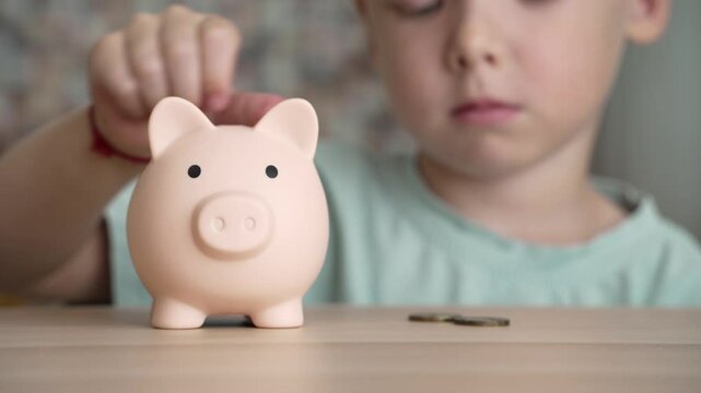 A young child carefully places a coin into a piggy bank. The child is focused on saving money.