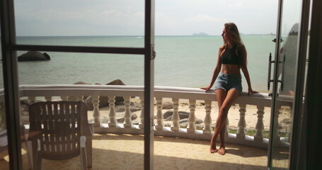 Obraz premium A woman is standing on a balcony next to the ocean, overlooking the waves crashing against the shore in from a resort in Thailand.