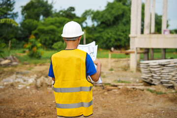 An architect, wearing a hardhat and safety vest, checks a laptop and house plan paper on a clipboard. Blueprints and construction documents lie nearby, emphasizing safety and precision in building.