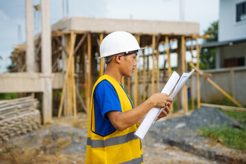 An architect, wearing a hardhat and safety vest, checks a laptop and house plan paper on a clipboard. Blueprints and construction documents lie nearby, emphasizing safety and precision in building.