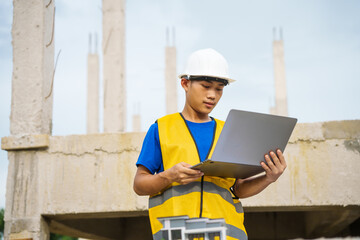 An architect, wearing a hardhat and safety vest, checks a laptop and house plan paper on a...
