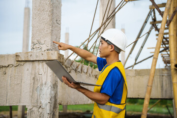 An architect, wearing a hardhat and safety vest, checks a laptop and house plan paper on a...