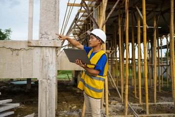 An architect, wearing a hardhat and safety vest, checks a laptop and house plan paper on a clipboard. Blueprints and construction documents lie nearby, emphasizing safety and precision in building.