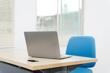 An empty desk in a minimalist office setting, featuring a blank workspace, large windows, a laptop, and a blue office chair against a white wall background, ideal for product montages. no people