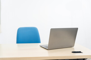 Fototapeta premium An empty desk in a minimalist office setting, featuring a blank workspace, large windows, a laptop, and a blue office chair against a white wall background, ideal for product montages. no people