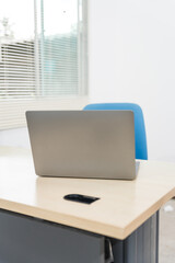 An empty desk in a minimalist office setting, featuring a blank workspace, large windows, a laptop, and a blue office chair against a white wall background, ideal for product montages. no people