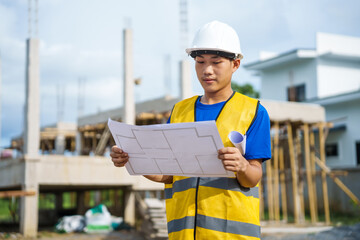 An architect, wearing a hardhat and safety vest, checks a laptop and house plan paper on a...