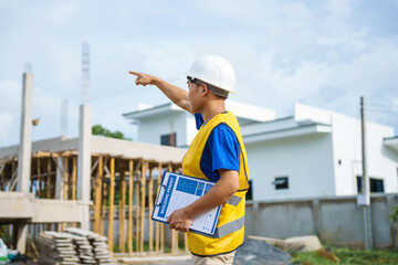 An architect, wearing a hardhat and safety vest, checks a laptop and house plan paper on a...