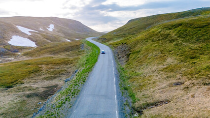A single car drives along a winding road through a green, hilly landscape in Norway. The road is flanked by lush, green vegetation and mountains in the distance. North Cape or Nordkapp, Norway