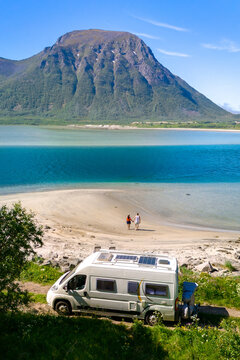 A couple walks hand-in-hand on a pristine beach, a campervan parked nearby, with a majestic fjord and mountain backdrop. motorhome at the beach of Lofoten Norway