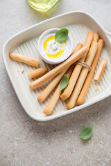 Serving tray with grissini bread sticks, ricotta dip and fresh basil, vertical shot on a beige stone background, elevated view