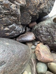 Close-up of an assortment of rocks and pebbles on a sandy beach, featuring various sizes and shapes with a smooth, polished texture. A large rock stands out among the surrounding smaller rocks.