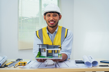 Asian architect works at a desk with house blueprints, checking plans on a laptop and clipboard,...
