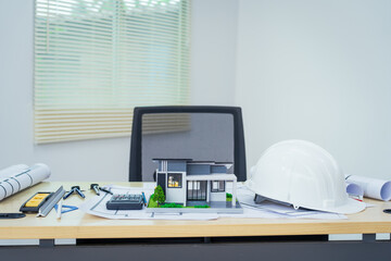 An architect's office with no people, featuring house blueprints, a model house toy, and architectural tools like a laptop, clipboard, hardhat, and safety vest for construction projects.