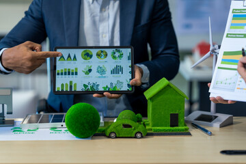 Fototapeta premium Two Asian businessmen in formal suits discuss ideas and suggestions at an office desk, focusing on green housing estates, city planning, renewable energy, and achieving net zero emissions by 2050.
