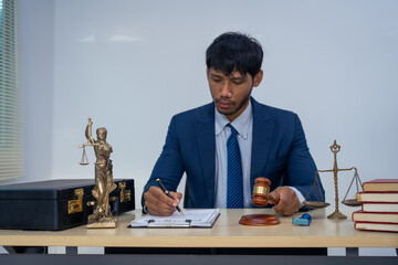An Asian businessman in a formal suit works alone at a lawyer's desk, by golden scales, law books,...