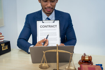 An Asian businessman in a formal suit works alone at a lawyer's desk, by golden scales, law books,...