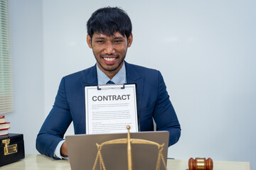 An Asian businessman in a formal suit works alone at a lawyer's desk, by golden scales, law books,...