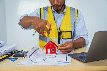Asian architect works at a desk with house blueprints, checking plans on a laptop and clipboard,...