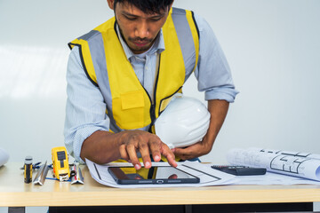 Asian architect works at a desk with house blueprints, checking plans on a laptop and clipboard,...