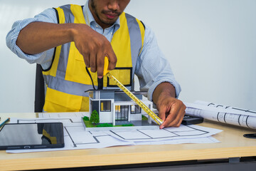 Asian architect works at a desk with house blueprints, checking plans on a laptop and clipboard,...