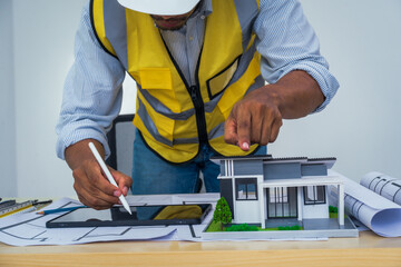 Asian architect works at a desk with house blueprints, checking plans on a laptop and clipboard,...