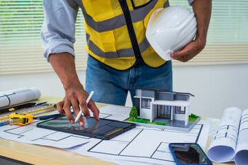 Asian architect works at a desk with house blueprints, checking plans on a laptop and clipboard,...