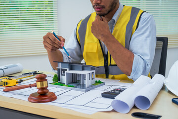 Asian architect works at a desk with house blueprints, checking plans on a laptop and clipboard,...