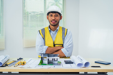 Asian architect works at a desk with house blueprints, checking plans on a laptop and clipboard,...