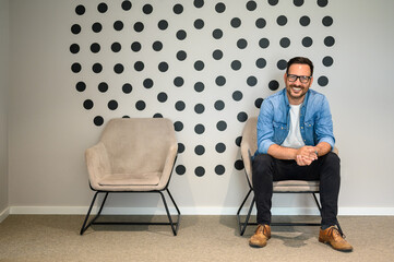 Portrait of handsome smiling freelancer in eyeglasses and with hands clasped sitting on office chair