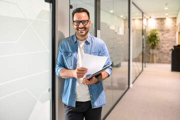 Portrait of confident male programmer with laptop and document standing by glass wall in modern office