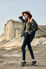 Stylish young woman in plaid shirt and jeans standing confidently in the desert wearing a hat