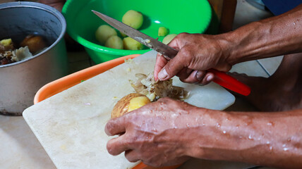 Close up of a person peeling potatoes with a knife in the kitchen. A hand peels a potato with a knife on a cutting board.