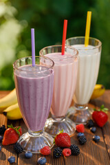 set of milkshakes in glasses with straw and fresh bananas and berries on table outdoors