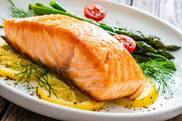 Fried salmon steak with cooked green asparagus, cherry tomatoes and lemon slices served on white plate on  wooden table