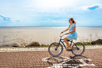 Fototapeta premium Active summer holidays. Woman with long hair wearing blue dress riding blue cruiser bike on bicycle path on seafront on sunny day. Side view