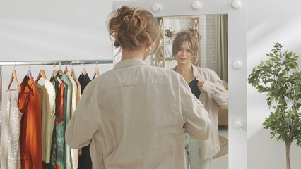 Young millennial woman in her bedroom looks in the mirror smiling, tries on a shirt, happy with...