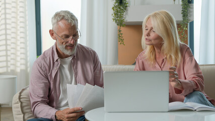 Fototapeta premium Happy mature modern Caucasian couple elderly senior female woman male man working laptop pc smile. Husband wife businesspeople analyzing documents papers discussing budget e-commerce online sofa home
