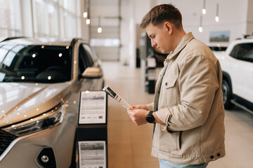 Side view of thoughtful young male customer holding papers, checking vehicle characteristics during...