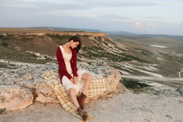 Woman sitting on top of a rock in front of a mountain with a plaid blanket enjoying a peaceful...