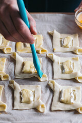 A hand's man brushing beaten egg on little puff pastry squares with camembert cheese pieces.