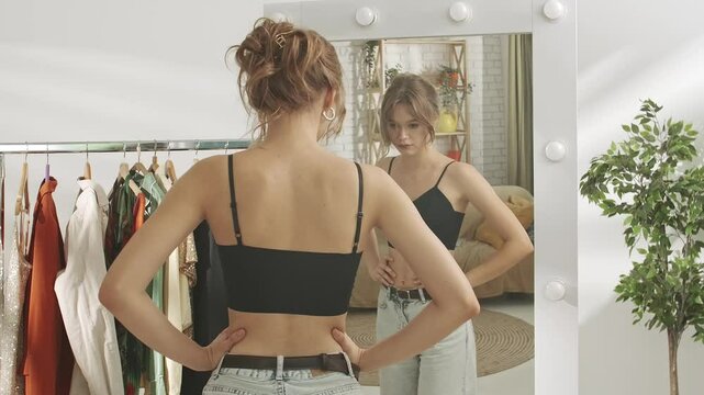 Young millennial woman in bedroom looks in the mirror, checks body appearance, not satisfied face self critics. Self care healthy beauty concept.