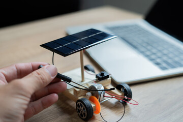 A person is building a solar powered toy car. The car is on a table next to a laptop