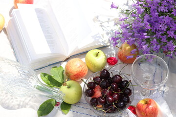 Open book, rose petals, apples, cherries and wine on a white tablecloth. Summer still life on a garden table. Leisure activities concept. 