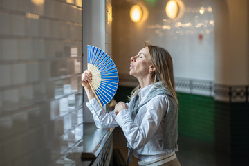 Exhausted girl suffering from stuffiness fanning, standing by window, breathing inhale fresh air. Distressed Businesswoman, female office worker left workplace to cope with sultriness in cabinet. 