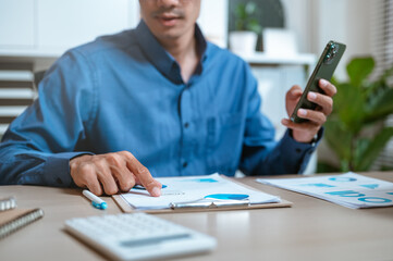 Fototapeta premium chart, document, graph, invoice, job, paperwork, planning, profit, report, smartphone. A man is sitting at a desk with a cell phone in his hand. He is looking at a piece of paper with a graph on it.