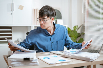 document, executive, job, manager, notice, papers, paperwork, read, stress, worried. A man in a blue shirt is sitting at a desk with a pile of papers in front of him. He is looking at the papers.